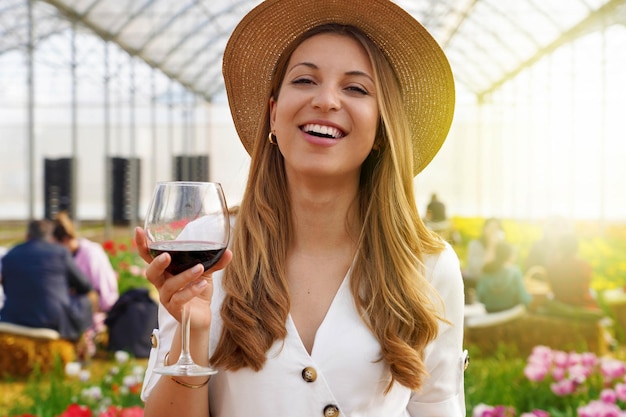 Mulher olhando para a câmera e segurando um copo de vinho com pessoas entre o campo de flores bebendo e conversando no fundo