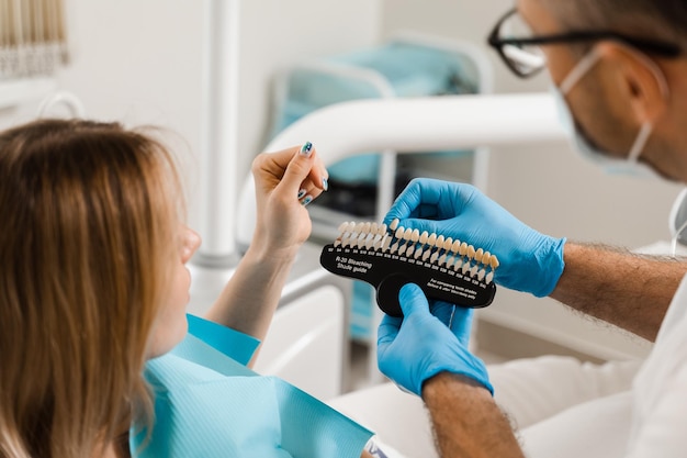 Mulher olhando para folheados ou implantes amostras de correspondência de cores dos dentes nas mãos do médico Odontologia O dentista demonstra o guia de tons de cor dos dentes para clareamento dental para paciente mulher na clínica odontológica