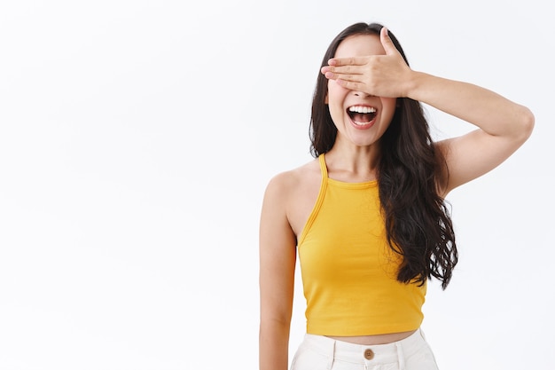 Mulher oriental alegre, feliz e despreocupada, cobrindo os olhos com a mão e sorrindo divertida, esperando pela surpresa do aniversário ou participando do concurso de festa de Halloween, sem espiar antecipando algo interessante