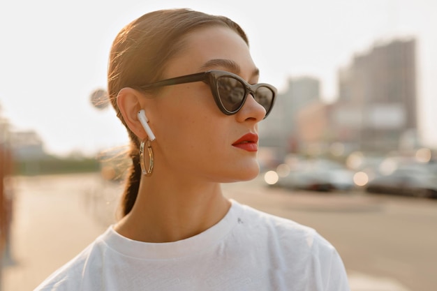 Mulher otimista com cabelo escuro em camiseta branca ouvindo música na cidade Moça legal na blusa clara e óculos escuros olhando de lado na cidade