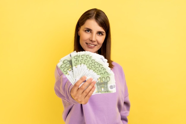Mulher otimista com cabelo segurando um grande fã de dinheiro olhando para a câmera com um sorriso