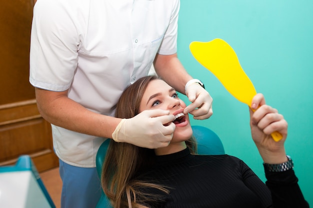 Mulher paciente com um sorriso lindo se senta em uma cadeira odontológica e olha no espelho o resultado