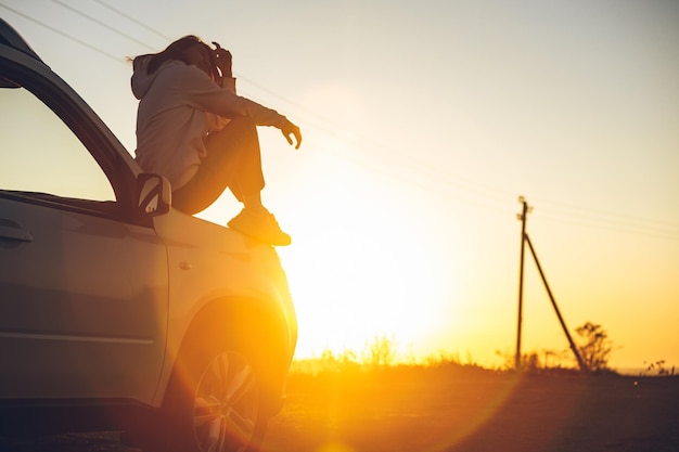 Mulher para na beira da estrada para apreciar o pôr do sol sentado no capô do carro