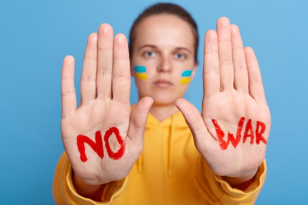 Mulher patriótica séria com capuz amarelo com bandeira ucraniana nas bochechas, mostrando as palmas das mãos sem inscrição de guerra posando isolada sobre fundo azul