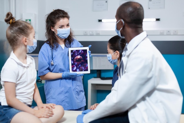 Mulher pediatra assistente com máscara de proteção contra coronavírus segurando computador tablet com experiência em vírus na tela. equipe médica discutindo os sintomas de covid19. serviço de saúde