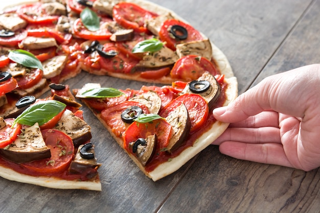 Mulher pegando pizza vegetariana com berinjela, tomate, azeitonas pretas, orégano e manjericão na superfície de madeira