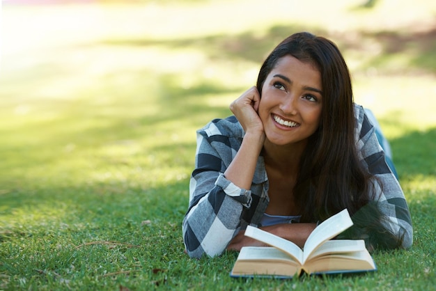 Mulher pensando e lendo livro sobre literatura de grama e sorrindo para ficção ou história de fantasia Pessoa feminina natureza e relaxando no gramado para conhecimento sonhando acordado e estudante estudando no jardim