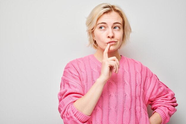 Mulher pensativa de camisola rosa com o dedo nos lábios olhando para longe em fundo claro