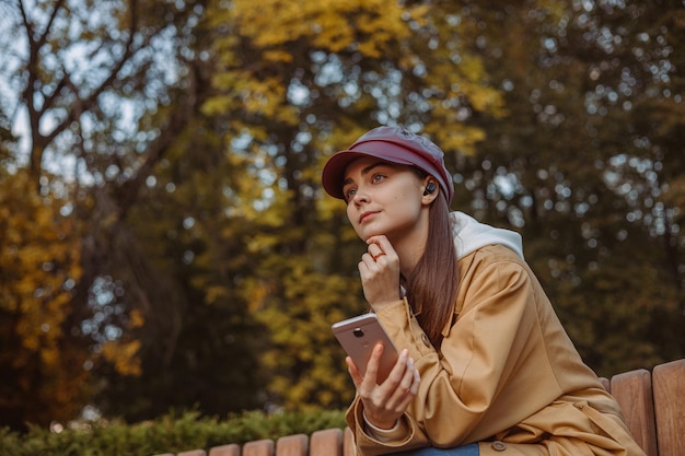 mulher pensativa em fones de ouvido sem fio com smartphone ouvindo música enquanto relaxa no parque