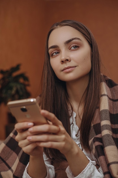 Mulher pensativa envolta em xadrez quente navegando smartphone e relaxando em casa