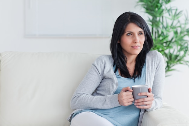 Mulher pensativa sentada em um sofá enquanto segura uma caneca