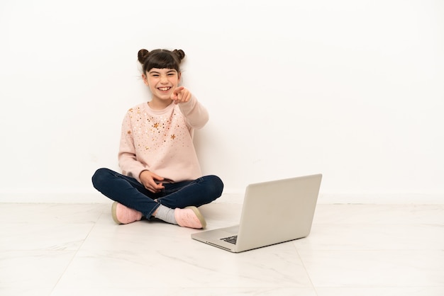 Mulher pequena com um laptop sentada no chão surpresa e apontando para a frente