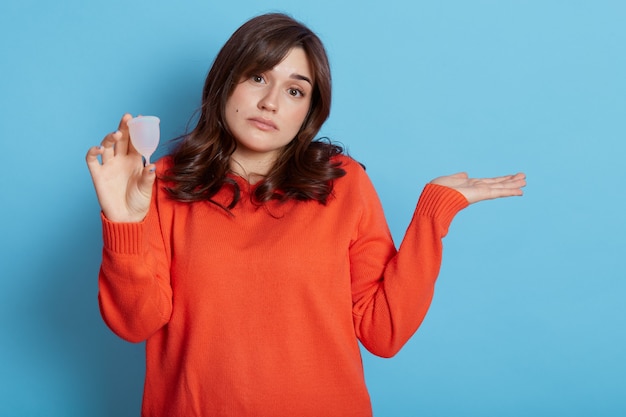 Mulher perplexa de cabelos escuros escolhe o produto durante os dias críticos, segura o copo menstrual