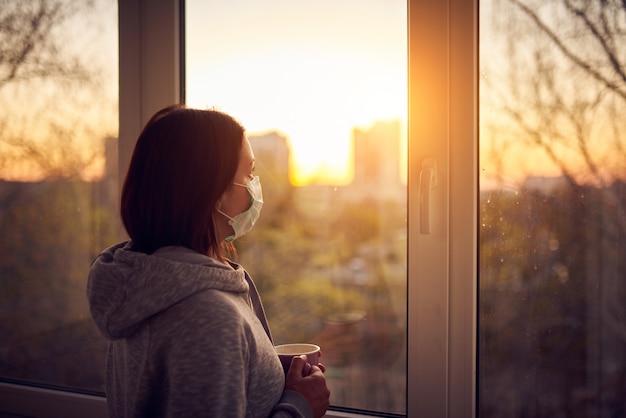 Foto mulher perto da janela ao pôr do sol em isolamento em casa para surto de vírus. ficar em casa conceito