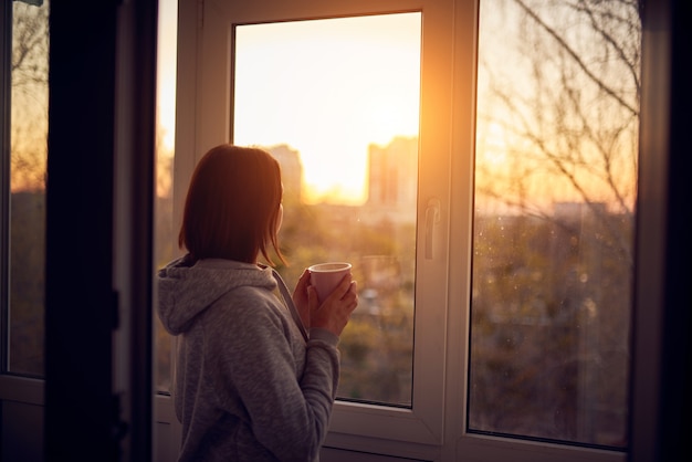 Mulher perto da janela ao pôr do sol em isolamento em casa para surto de vírus. Ficar em casa conceito