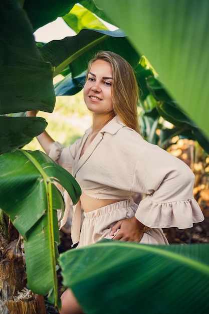 Mulher perto de folhas verdes de arbustos de banana na natureza em um parque em um lugar tropical ela está em uma saia bege e uma blusa