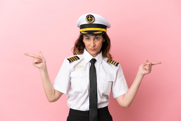 Mulher piloto de avião de meia-idade isolada em fundo rosa apontando o dedo para as laterais e feliz