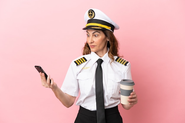 Mulher piloto de avião de meia-idade isolada em um fundo rosa segurando um café para levar e um celular