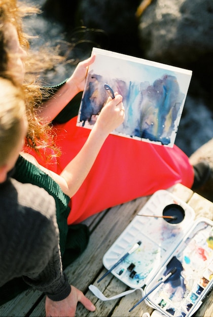 Foto mulher pintando um quadro enquanto está sentada ao lado de um homem em uma ponte de madeira