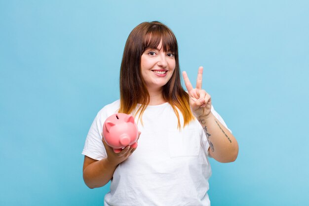 Mulher plus size sorrindo e parecendo feliz, despreocupada e positiva, gesticulando vitória ou paz com uma mão