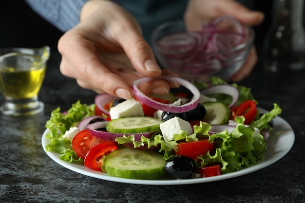 Mulher põe cebola na salada grega na mesa preta esfumada