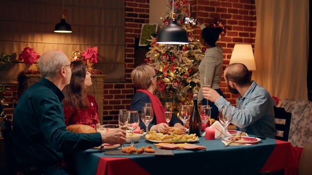 Mulher positiva ao lado da árvore de natal enquanto tira foto de selfie de grupo familiar no jantar. parentes festivos se reuniram em torno da mesa de jantar enquanto celebravam juntos o feriado de inverno.