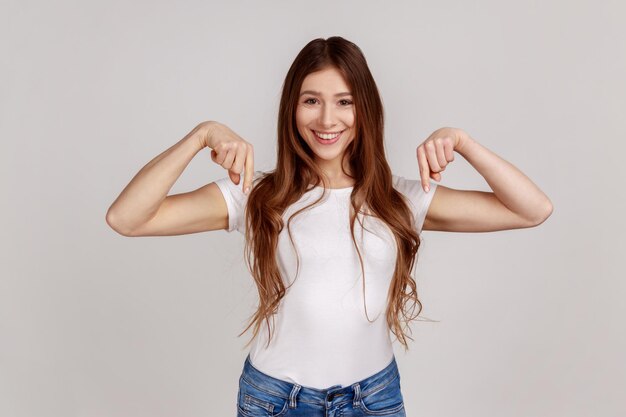 Mulher positiva, apontando os dedos para baixo, mostrando espaço para o seu anúncio, olhando para a câmera com sorriso, vestindo camiseta branca, tiro de estúdio interno isolado em fundo cinza