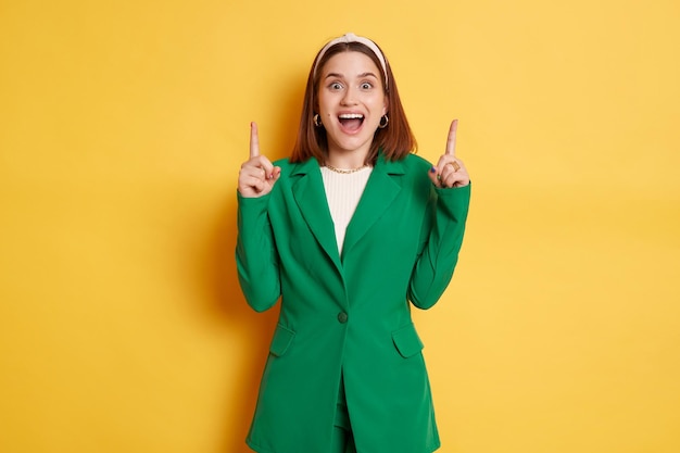 Mulher positiva atônita vestindo jaqueta verde isolada sobre fundo amarelo apontando para cima no espaço vazio da cópia da parede em branco para propaganda