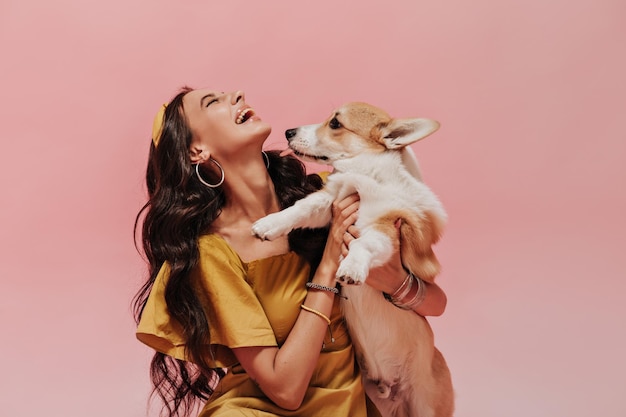 Mulher positiva com cabelo comprido em brincos e bandana em roupa amarela da moda rindo e segurando cachorro fofo no pano de fundo rosa