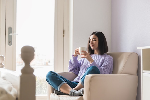 Mulher positiva de etnia atraente jovem em seu quarto segurando e desfrutando de uma xícara de café.