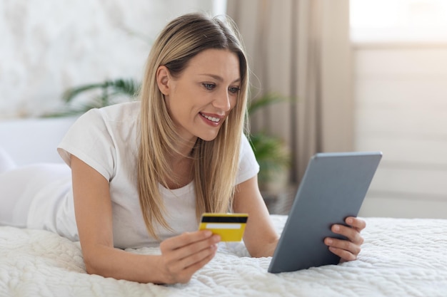 Mulher positiva deitada na cama com tablet e cartão de crédito