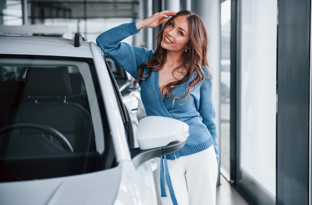 Mulher positiva na camisa azul, apoiando-se no carro novo no salão de automóveis