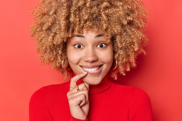 Mulher positiva olha com interesse para a câmera morde os lábios reage em algo bom veste casual gola alta com olhos cheios de felicidade na câmera isolada sobre fundo vermelho. emoções felizes