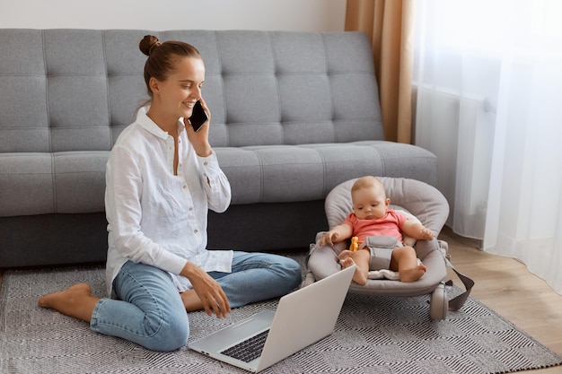 Mulher positiva sorridente na camiseta branca sentada perto da tosse com a filha bebê na cadeira de balanço e trabalhando no laptop falando no celular de bom humor