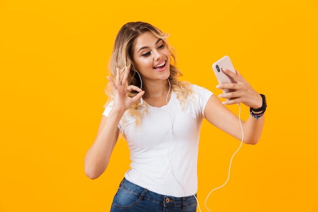 Mulher positiva usando fones de ouvido sorrindo e tirando foto de selfie no celular, isolada sobre a parede amarela