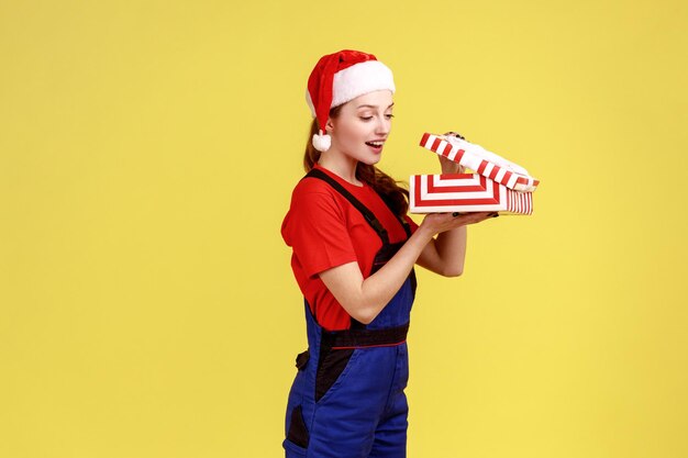 Mulher prática de pé com caixa de presente aberta, olhando para dentro com sorriso e interesse no interior, vestindo macacão azul e chapéu de papai noel. Tiro de estúdio interior isolado em fundo amarelo.