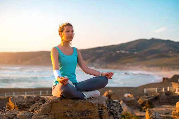 Mulher pratica ioga pose de lótus para meditação com férias de verão praia rochosa felicidade e relaxamento Exercício feminino calmo com ioga meditar praia oceânica com tempo dourado do pôr do sol