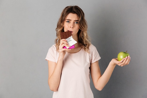 Mulher preocupada em t-shirt, comer chocolate, mantendo a maçã fresca e cinza