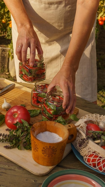 mulher preserva tomates secos em potes com alho picado e manjericão fresco no jardim ao pôr do sol