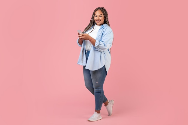 Mulher preta plus size usando smartphone em pé no fundo rosa