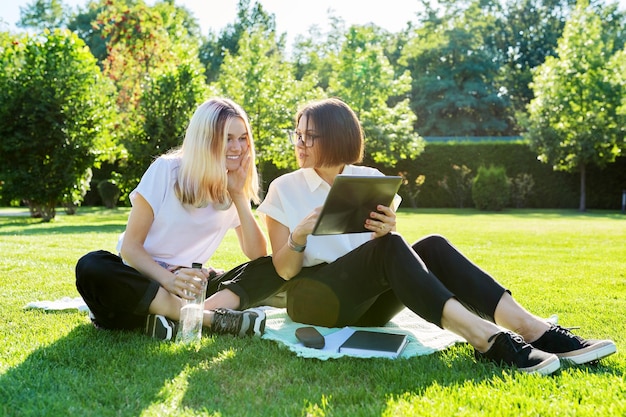 Mulher professora psicóloga assistente social conversando com uma estudante adolescente sentada na grama no parque da faculdade falando usando tablet digital