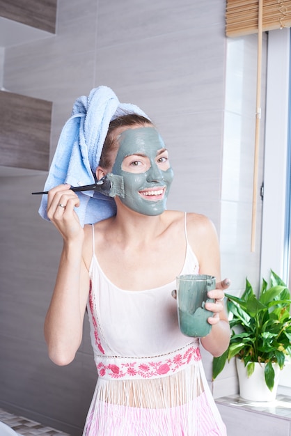 Mulher que aplica o creme de pele hidratando da máscara no rosto que olha no espelho do banheiro. Menina cuidando de seu creme hidratante de camadas de pele. Tratamento de spa para a pele.