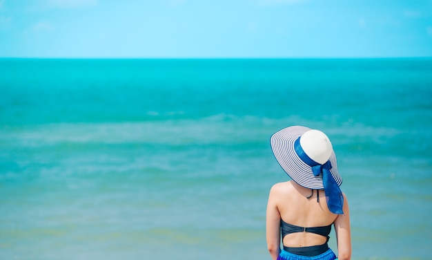 Mulher que aprecia a praia que relaxa alegre no verão pela água azul tropical. Modelo de biquíni na viagem usando chapéu de praia.