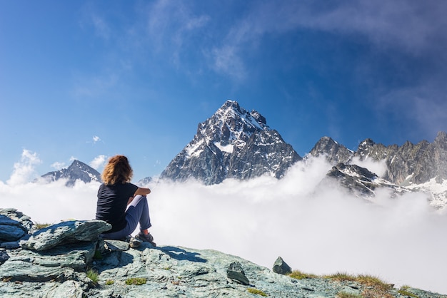 Mulher que descansa no topo da montanha alta, olhando o pico de montanha majestoso sobre as nuvens da paisagem dramática da vista, céu azul claro.