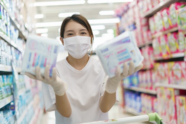 Mulher que desgasta a máscara cirúrgica e as luvas, comprando o guardanapo sanitário no supermercado. Compras de pânico após pandemia de coronavírus.