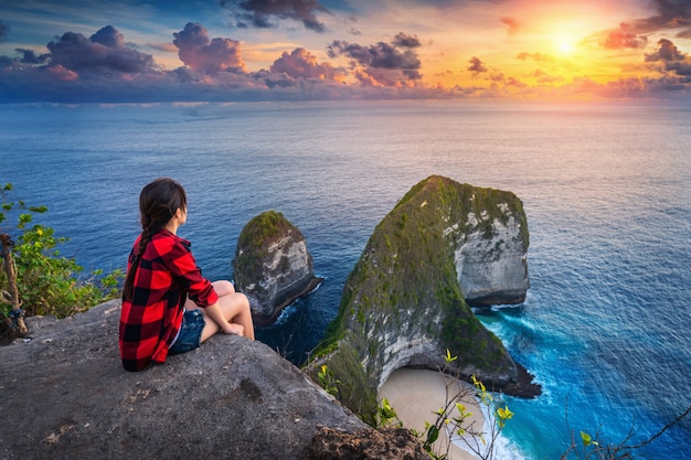 Mulher que senta-se no penhasco e que olha o por do sol na praia de Kelingking na ilha de Nusa penida, Bali, Indonésia.