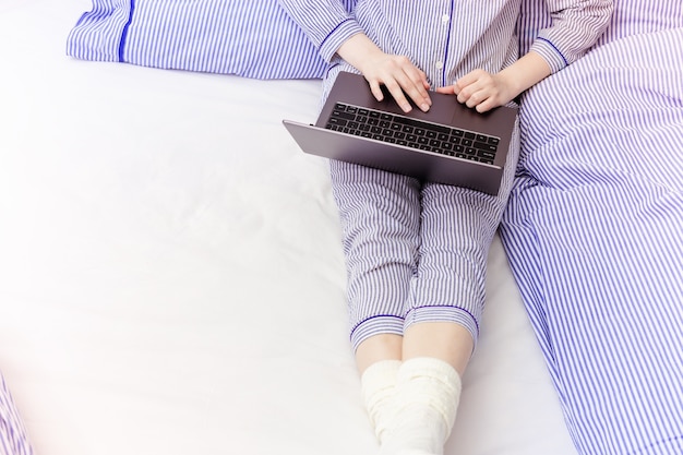 Mulher que trabalha em casa com o laptop de pijama e sentada na cama.