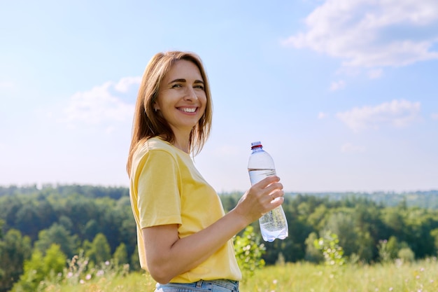 Mulher quente e ensolarada de verão com garrafa de água olhando para a câmera para nós natureza