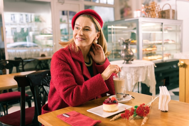 Mulher radiante. Mulher madura e elegante tomando café com leite e comendo sobremesa em uma confeitaria aconchegante