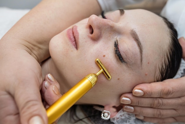 Mulher recebe massagem facial de pulso no spa vibrando massageador facial dourado no rosto feminino